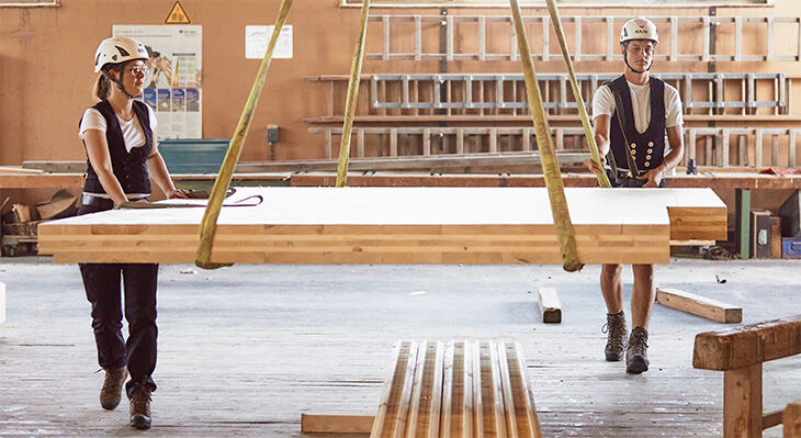 Eine Zimmerin und ein Zimmerer arbeiten in einer Werkstatt und bewegen gemeinsam ein Holzbauelement