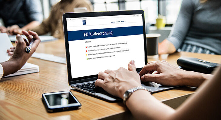 Mehrere Personen sitzen an einem Tisch. Im Vordergrund arbeitet eine Frau an ihrem Laptop und hat eine Webseite zur EU KI-Verordnung geöffnet.