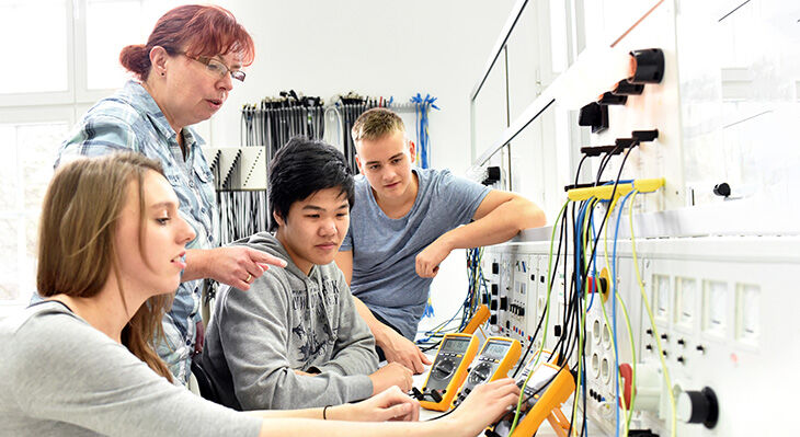 Das Bild zeigt drei Jugendliche, die unter Anweisung einer Ausbilderin Strommessungen durchf&uuml;hren.